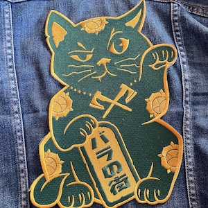 Op de afbeelding: Geborduurde patch met een groene Maneki-neko kat met gouden accenten, die een gouden tablet met Japanse tekst vasthoudt. De patch is op een spijkerjas genaaid. De kat heeft een opgeheven poot en bloemmotieven.