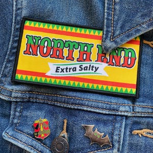 Op de afbeelding: Een spijkerjasje met een rechthoekige patch met de tekst "NORTH END Extra Salty" in rode en groene letters op een gele achtergrond. Onder de patch zitten drie pins: een schild, een Levi Strauss pin en een vleermuisvormige pin.