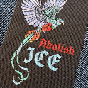 Peut inclure: Un écusson rectangulaire noir avec un oiseau coloré aux ailes déployées. L'oiseau présente des détails bleus, verts, rouges et oranges. Le texte "Abolish ICE" est en rouge et "ICE" en bleu clair.