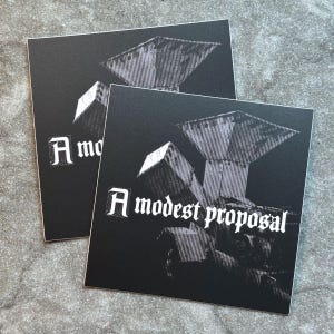 May include: Two square black cards with the text "A modest proposal" in white gothic font. The cards feature a detailed grayscale illustration of a structure with a geometric design. The cards are on a gray surface.