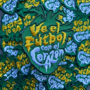 Op de afbeelding: Een verzameling groene, geborduurde patches met een hartvorm. De patches tonen de tekst "Ve el Fútbol con el Corazón" in geel en wit. De patches zijn waarschijnlijk bedoeld voor kleding of accessoires.