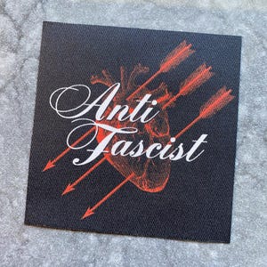 May include: Square black patch with the words "Anti Fascist" in white script, with a red heart pierced by three red arrows. The patch has a textured appearance.