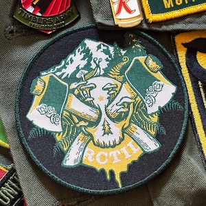 May include: A round, embroidered patch with a skull and crossed axes design. The patch features green, white, and yellow colors, with the word "RCTIL" in yellow. The patch is surrounded by other patches and pins.