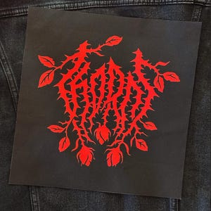 May include: A square black fabric patch featuring a bright red graphic design of thorny vines and rosebuds. The central text is stylized in a jagged, sharp font that mimics the appearance of thorns, integrated into the botanical illustration.