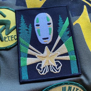 Op de afbeelding: Geborduurde patch met een lichtblauw gezicht karakter met een wit masker ontwerp. De patch heeft een zwarte achtergrond met groene en gele accenten, en een sterrenburst ontwerp.
