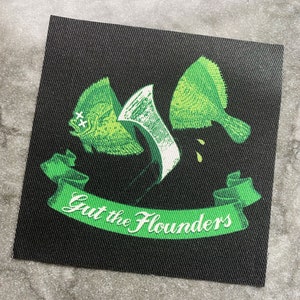 Op de afbeelding: Vierkante zwarte patch met een groen-witte illustratie van een vis die wordt opengesneden. De tekst "Gut the Flounders" staat op een groene banner eronder.