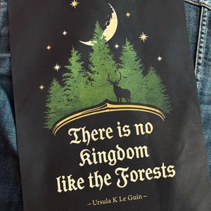 Könnte beinhalten: Ein schwarzer rechteckiger Druck mit einer grünen Waldsilhouette, einem Hirsch, einer Mondsichel und Sternen. Der Text lautet: There is no kingdom like the Forests - Ursula K Le Guin - in einer dekorativen goldenen Schrift unter der Grafik.