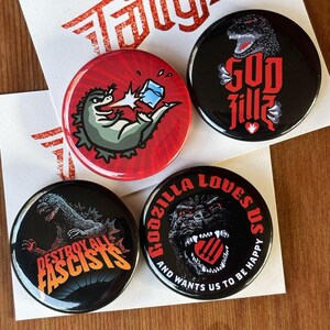 May include: Four round pinback buttons with Godzilla-themed designs. One button features Godzilla with the text "GODZILLA". Another button has Godzilla with the text "DESTROY ALL FASCISTS". The third button shows Godzilla with an ice cube. The last button says "GODZILLA LOVES US".
