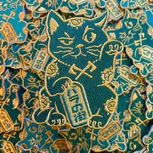 Op de afbeelding: Teal en goud geborduurde patch met een gestileerde Maneki-neko kat met een opgeheven poot. De kat houdt een bord met Japanse tekst vast. De achtergrond is gevuld met vergelijkbare patches.