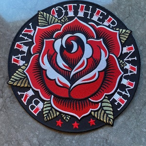 May include: Embroidered patch featuring a red rose with white and black details, surrounded by the text "BY ANY OTHER NAME" in white. The patch has gold and green leaves and three red stars on a black background.