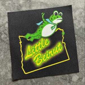 May include: Black square patch featuring a green frog with a blue scarf, above a yellow outline of a state with the words "Little Beirut" in yellow and green. The patch has a textured surface.