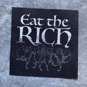 Puede incluir: Un parche cuadrado negro con texto blanco que dice "Eat the RICH" en una fuente gótica. Debajo del texto hay una ilustración estilizada y descolorida. El parche está hecho de una tela texturizada.