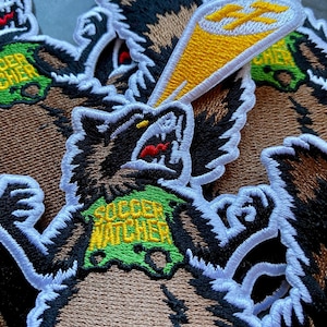 Op de afbeelding: Geborduurde patch met een cartoon das die een biertje vasthoudt, met de tekst "SOCCER WATCHER" op een groene achtergrond. De das heeft bruine vacht, zwart-witte details en een rode mond. De patch heeft een witte rand.