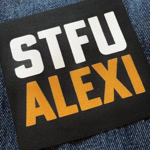 May include: A square black fabric patch featuring the white text STFU and the yellow text ALEXI printed in a bold, sans-serif font. The patch has a visible woven texture and is positioned against a dark blue denim fabric background.