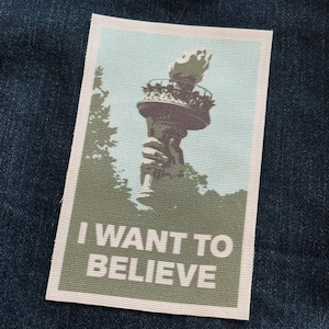 May include: A rectangular patch featuring the Statue of Liberty in shades of green and brown against a light blue sky. The text "I WANT TO BELIEVE" is printed in white block letters below the image. The patch is set against a dark blue denim background.