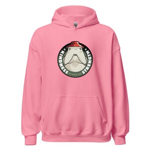 Op de afbeelding: Een roze hoodie met een cirkelvormige afbeelding van een cartoon walrus met een hoed. De afbeelding leest "Kindness Gives Us Strength" in een cirkel rond de walrus.