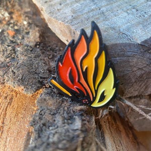 May include: A colorful enamel pin in the shape of a flame. The pin features a black outline with vibrant colors including red, orange, yellow, and a small amount of white. The pin is set against a textured wooden surface.