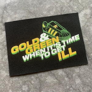 Op de afbeelding: Zwart rechthoekig embleem met de tekst "GOLD & GREEN WHEN IT'S TIME TO GET ILL" in goud en groene letters. Een groene en gouden sneaker-afbeelding staat boven de tekst. Het embleem is gemaakt van een textuurstof.
