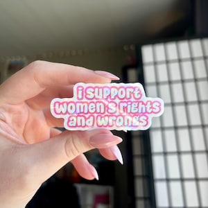 Puede incluir: Una pegatina holográfica con el texto "i SUPPORT WOMEN'S RIGHTS AND WRONGS" en rosa y blanco. La pegatina se sostiene en una mano con esmalte de uñas rosa. El fondo está borroso.
