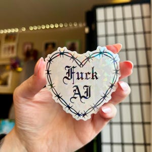 Könnte beinhalten: Ein herzförmiger Aufkleber mit holografischem Finish. Der Aufkleber zeigt den Text "Fuck AJ" in einer gotischen Schrift, umgeben von einem Stacheldrahtdesign. Der Aufkleber wird in einer Hand gehalten.