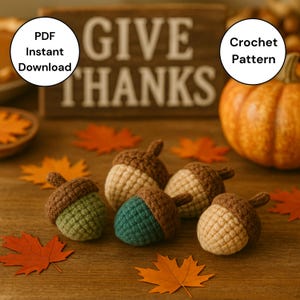 May include: Five crocheted acorns in brown, green, and beige sit on a wooden surface with autumn leaves. A wooden sign saying "GIVE THANKS" and a pumpkin are in the background. The image also includes text that says "PDF Instant Download" and "Crochet Pattern".