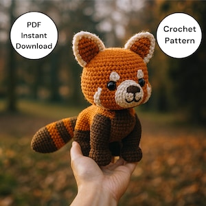 May include: A handmade crocheted red panda toy, featuring a round head, large ears, and a striped tail. The toy is mainly orange with brown detailing. The image includes text that says "PDF Instant Download" and "Crochet Pattern".