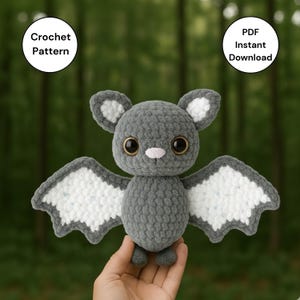 Pode incluir: Um morcego de crochê cinza feito à mão com asas brancas e cinzas, olhos grandes e pretos e um nariz rosa. O morcego é segurado em uma mão. A imagem inclui o texto "Crochet Pattern" e "PDF Instant Download".