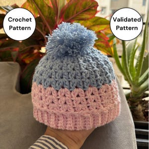 May include: A handmade crochet hat in light blue and pink, topped with a blue pom-pom. The hat features a textured stitch pattern and is designed for warmth and style. The text "Crochet Pattern" and "Validated Pattern" are in the image.