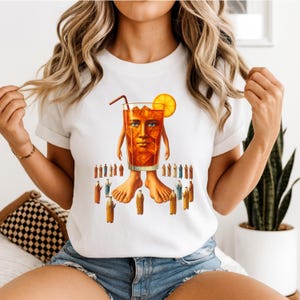 May include: White t-shirt with a surreal illustration of a drink with a human face, arms, and legs. The drink has a straw and orange slice. Small figures in robes surround the drink.