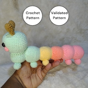 May include: A handmade crochet caterpillar toy in pastel colors. The caterpillar has a light green head, a pink mouth, and a series of body segments in light green, yellow, peach, and pink. The text "Crochet Pattern" and "Validated Pattern" are in the background.