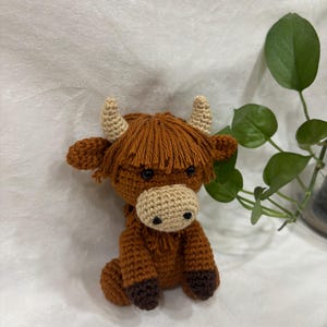May include: A crocheted Highland cow figurine in shades of brown, tan, and beige. The handmade cow has a shaggy mane, small horns, and a light-colored muzzle. The cow is sitting with its legs tucked under its body.