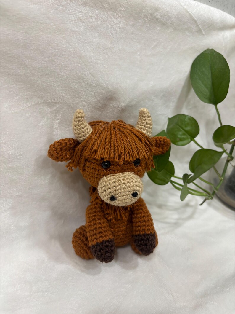 May include: A crocheted Highland cow figurine in shades of brown, tan, and beige. The handmade cow has a shaggy mane, small horns, and a light-colored muzzle. The cow is sitting with its legs tucked under its body.