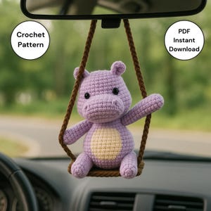 May include: A crocheted purple hippo toy hanging from a brown rope swing. The hippo has a light yellow belly and black eyes. Text on the image reads "Crochet Pattern" and "PDF Instant Download".