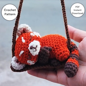 Może przedstawiać: Szydełkowa zabawka w kształcie czerwonej pandy z białymi i brązowymi akcentami, zawieszona na brązowej nici. Panda leży na małej, szydełkowej brązowej huśtawce. Tekst na obrazku to "Crochet Pattern" i "PDF Instant Download".