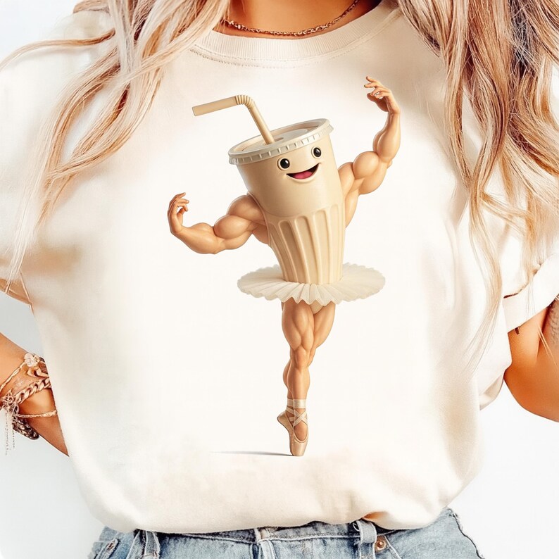 May include: A cream-coloured t-shirt with a playful illustration of a milkshake character. The milkshake has muscular arms and legs, a smiling face, and wears a white tutu and ballet shoes. A straw extends from the top of the milkshake cup.