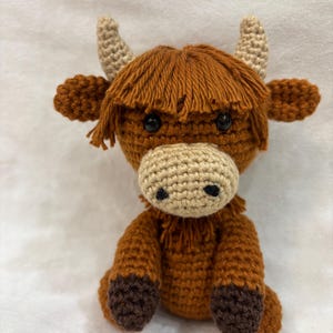May include: A handmade crochet Highland cow plushie. The plushie features a brown body, head, and legs with a lighter tan muzzle and horns. The cow has shaggy brown hair and black eyes. The toy is sitting upright.