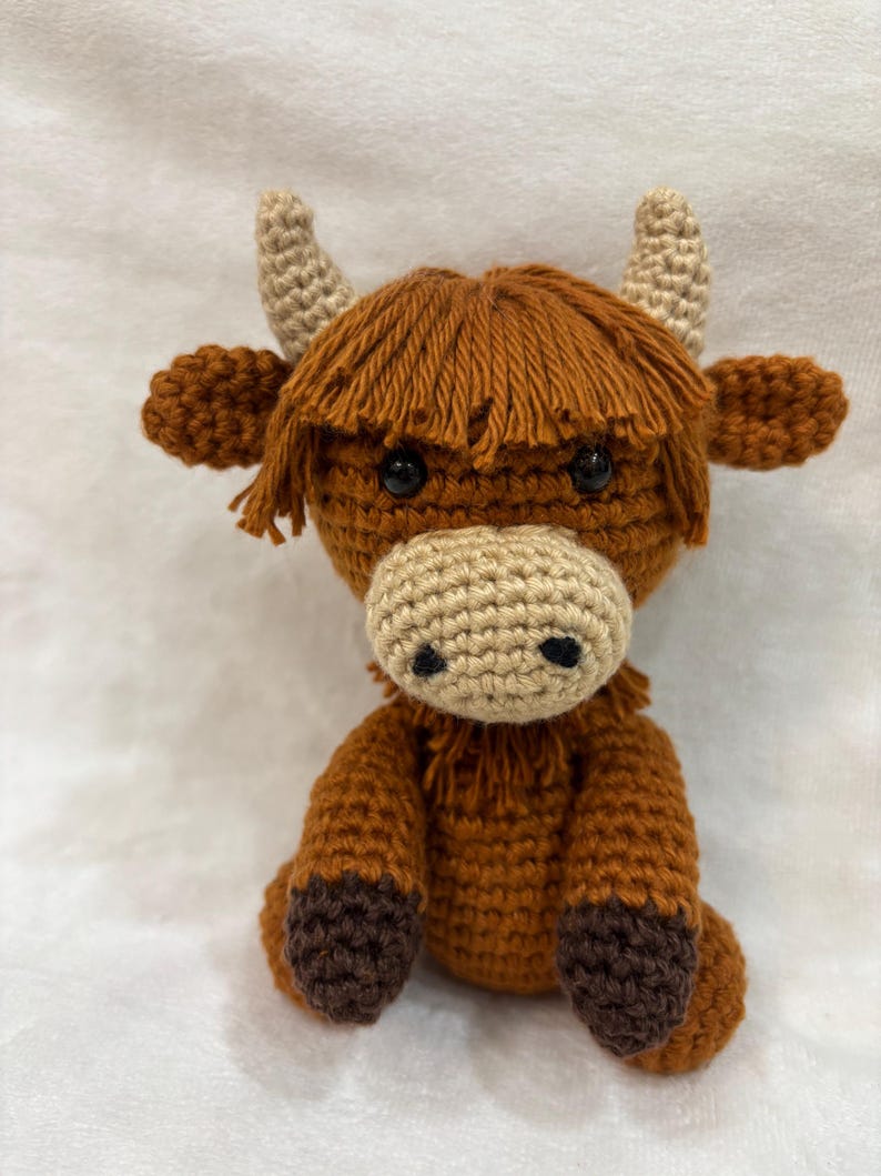 May include: A handmade crochet Highland cow plushie. The plushie features a brown body, head, and legs with a lighter tan muzzle and horns. The cow has shaggy brown hair and black eyes. The toy is sitting upright.
