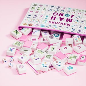 May include: A Mahjong set featuring white tiles with colorful floral, bamboo, and character designs, along with three white dice. The tiles are scattered in front of a white zippered storage case printed with the text MAH JONG SEWN in purple.