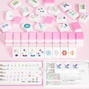May include: A set of white mahjong tiles with pink backs, featuring colorful floral and bamboo designs. The set includes a row of tiles with flower illustrations, a reference card with game rules, and two small white dice on a pink surface.