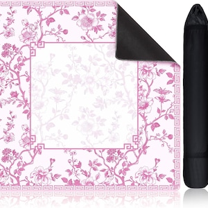May include: A square mat featuring a pink and white floral pattern with a geometric border and a central blank rectangular space. The mat has a black underside and is shown alongside a black cylindrical carrying case with a shoulder strap.