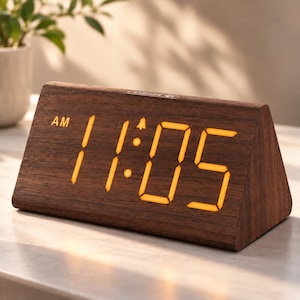 May include: A triangular digital alarm clock with a dark brown wood-grain finish. The front face displays the time 11:05 in glowing orange LED numbers, with an AM indicator and a small bell icon. The word SNOOZE is printed on the top edge.