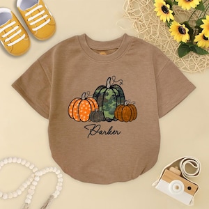 May include: A brown baby bodysuit featuring a pumpkin graphic. The graphic includes an orange pumpkin with white polka dots, a green camouflage pumpkin, a small gray pumpkin, and a brown pumpkin. The name "Parker" is written in script below the pumpkins.