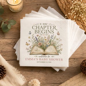 May include: A stack of white paper napkins featuring a printed illustration of an open book with a bouquet of wildflowers and butterflies. The text reads A NEW CHAPTER BEGINS, welcome to EMMA'S BABY SHOWER, and SEPTEMBER 10, 2026 in dark ink.