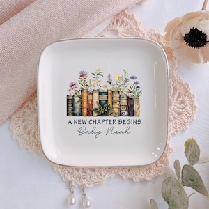 May include: A white square ceramic trinket dish with a gold rim. The dish features a design of vintage books with colorful flowers and the text "A New Chapter Begins Baby Noah".
