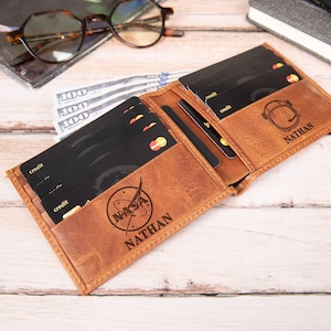 May include: Open brown leather wallet with multiple black credit cards and cash. The wallet has the NASA logo and the name "NATHAN" engraved on it. The wallet is open, revealing multiple card slots and a cash compartment.