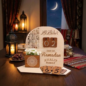 May include: Wooden Ramadan calendar with a decorative floral box and date tiles. The calendar features the text "Day of Ramadan" and "Khalif Family". The background includes a crescent moon and lanterns.