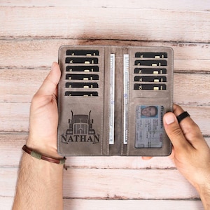 May include: Gray leather wallet with multiple card slots, cash compartments, and an ID window. The wallet features a truck graphic and the name "NATHAN" engraved on the front. The wallet is open, revealing its interior.
