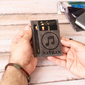 May include: Gray leather cardholder with a music note design and the name "NATHAN" engraved below. The cardholder is filled with credit cards. A brown and green bracelet is visible in the foreground.
