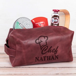 May include: A burgundy leather toiletry bag with a chef's hat and the name "Chef Nathan" engraved on the front. The bag is filled with grooming products, including a hair comb, hair styling product, and a bottle of aftershave.