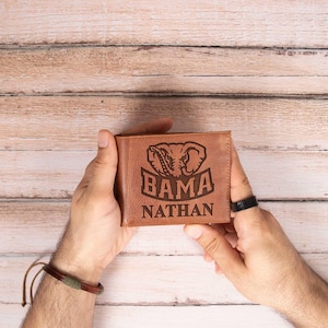 May include: A brown leather wallet with the word "BAMA" and an elephant logo engraved on the front. The name "NATHAN" is also engraved below the logo. The wallet is held by a person.